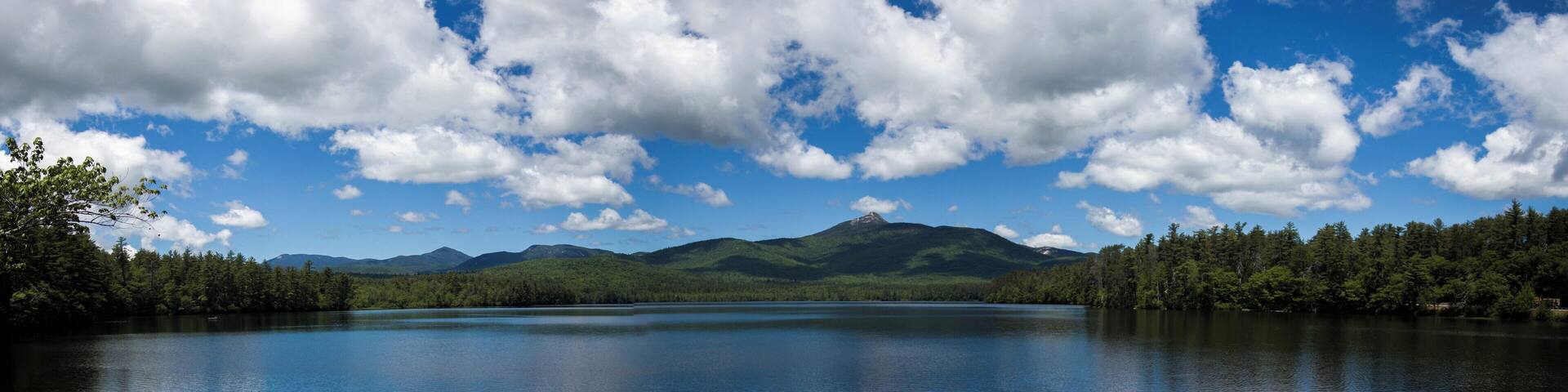 A calm lake surrounded by dense forests and mountains under a partly cloudy sky.