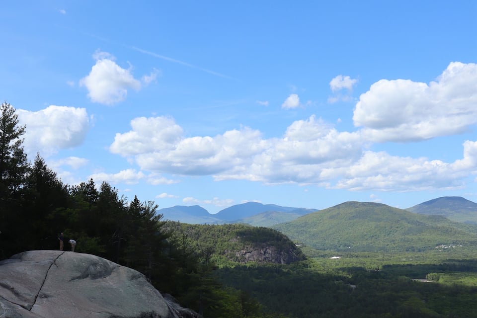 A rocky outcrop with a person standing on it, overlooking a forested valley with mountains in the distance and a clear blue sky.