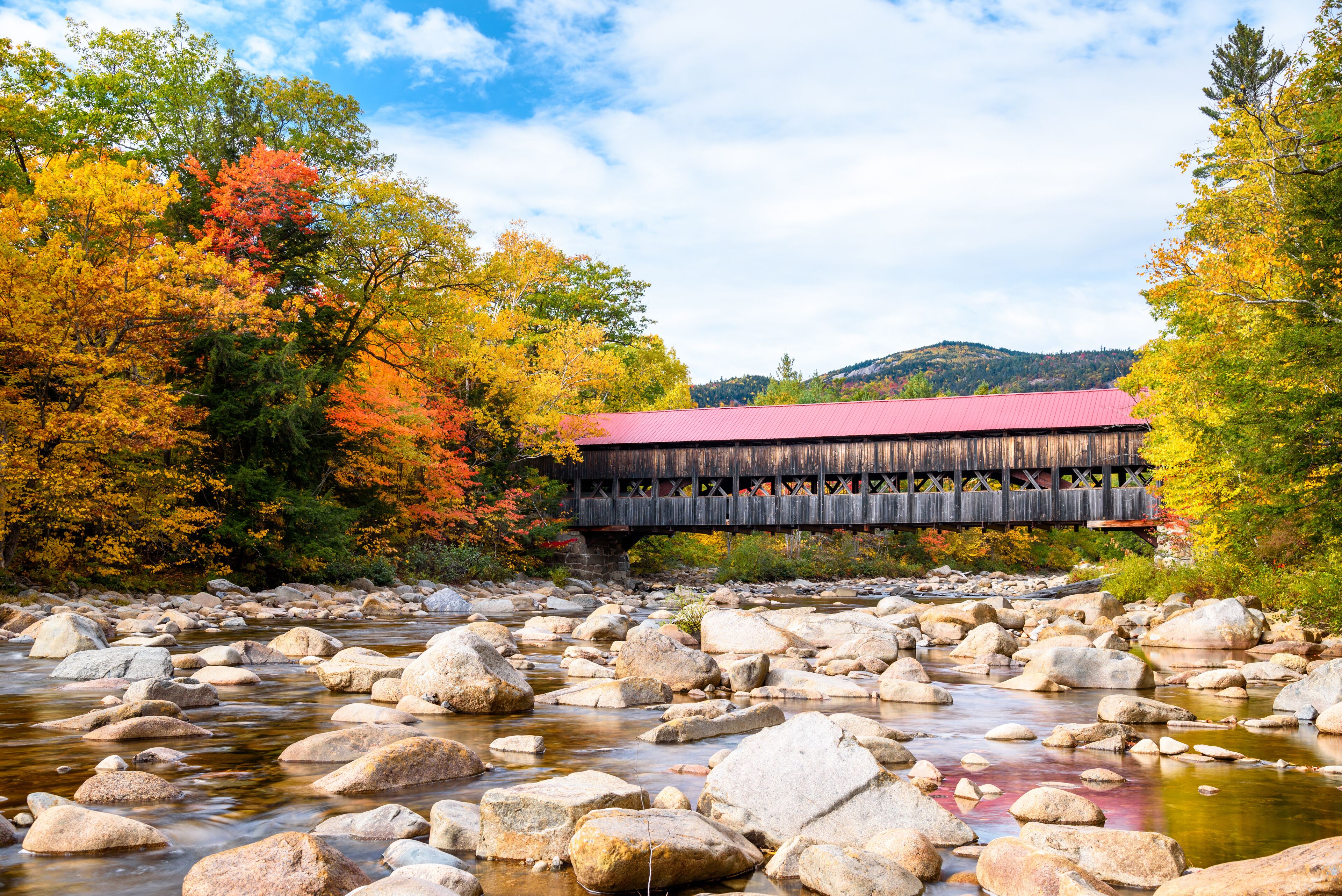 Historic covered bridge spanning a mountain river running through a colourful forest at the peak of fall foliage on a partly cloudy autumn day. Stunning autumn colours.