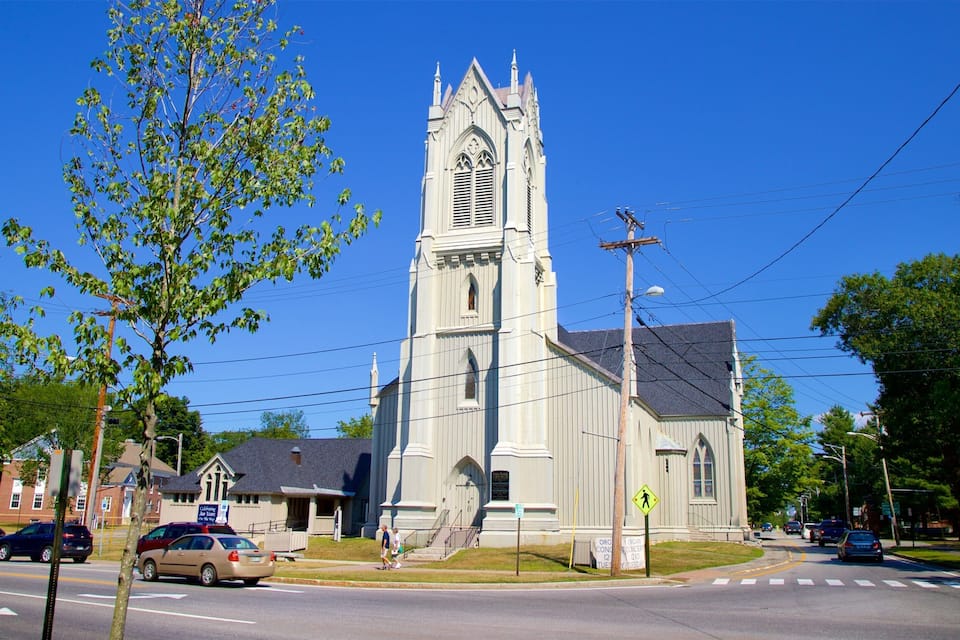 A church with a tall steeple and a cross on top, surrounded by trees and a street with cars.
