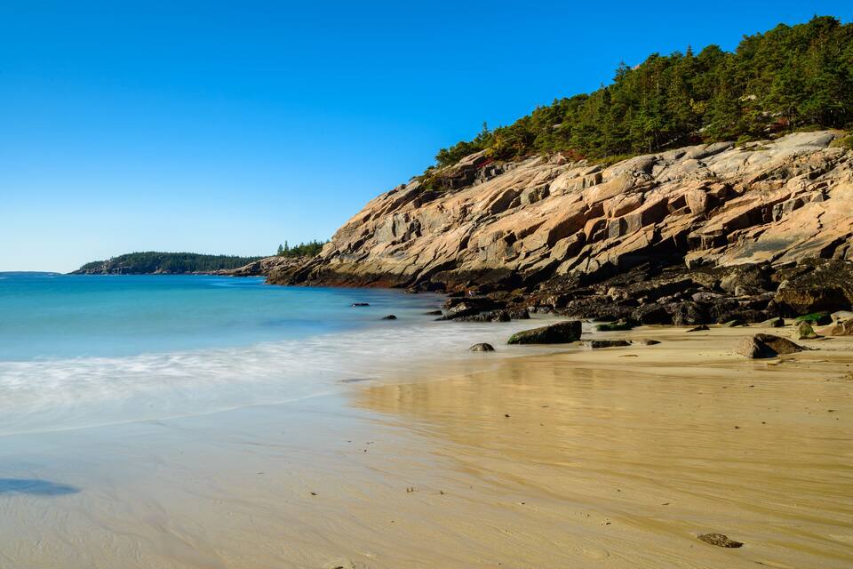 A beach with clear blue water, sandy shore, and rocky cliffs with trees.