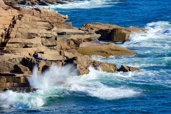 Une côte rocheuse où les vagues s’écrasent contre les rochers, et une personne qui prend une photo.