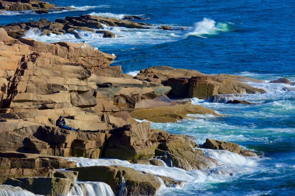 Une côte rocheuse où les vagues s’écrasent contre les rochers, et une personne assise sur ces rochers.
