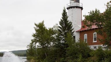 A lighthouse with a red roof and white walls, surrounded by trees and rocky shoreline.