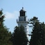 A lighthouse with a white tower and a dark top, surrounded by trees.