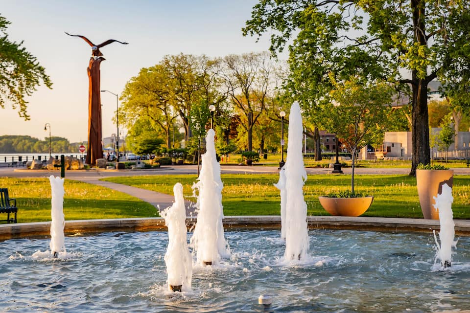 A fountain with multiple jets of water, surrounded by trees and a tall sculpture.