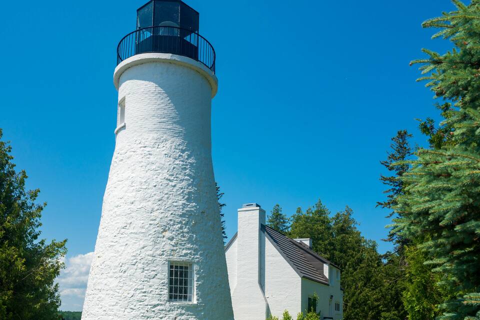 A white lighthouse with a black top, a white house, and green trees in the background.