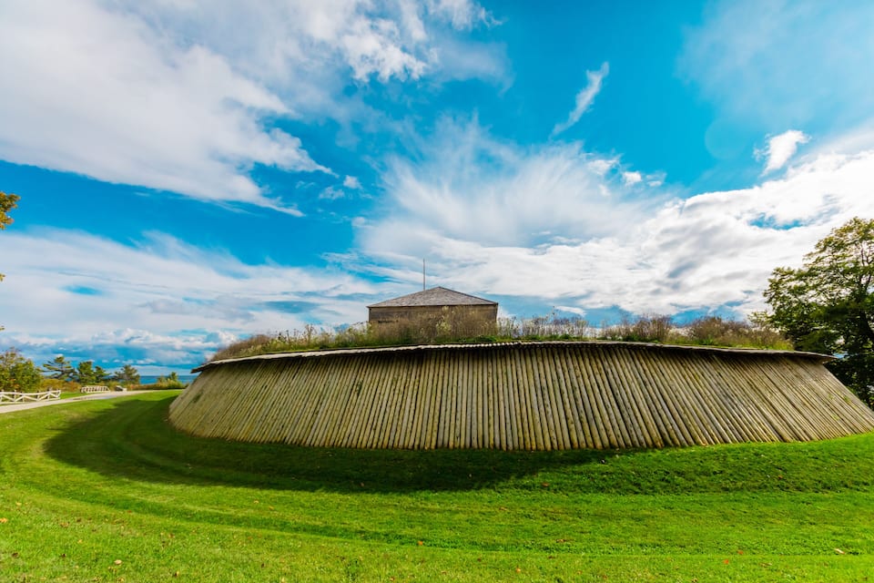 A large earthen mound with a small building on top, surrounded by green grass and trees under a blue sky with clouds.
