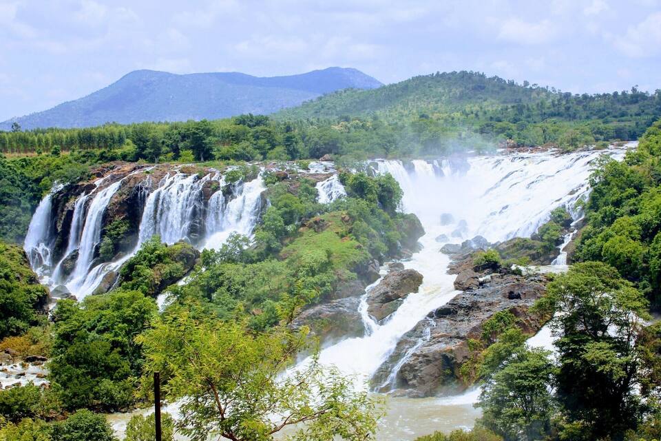 A multi-tiered waterfall surrounded by lush greenery and mountains in the background.
