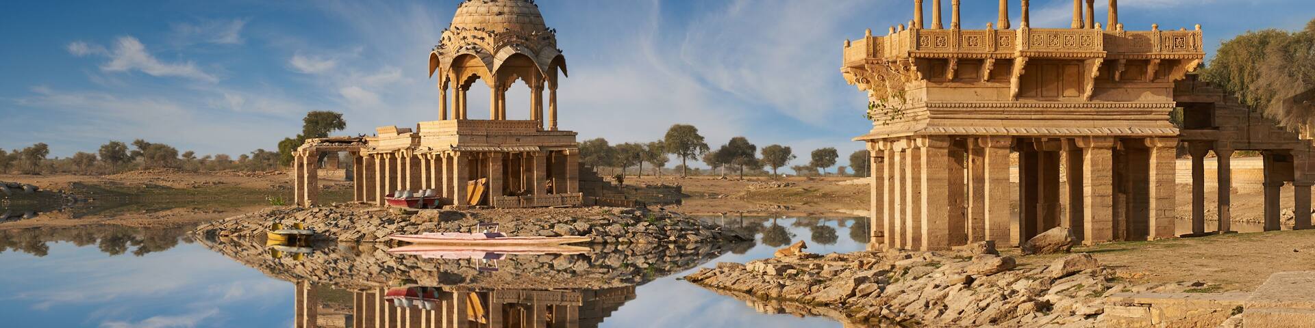 A calm lake reflecting two ornate domed structures with intricate carvings, surrounded by a rocky landscape and sparse trees.