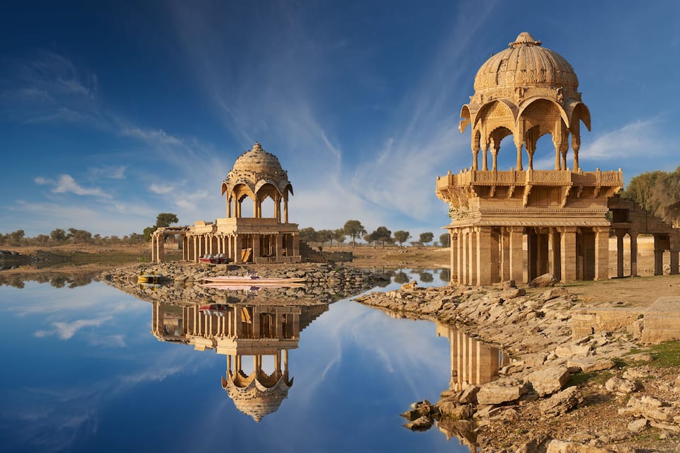 A calm lake reflecting two ornate domed structures with intricate carvings, surrounded by a rocky landscape and sparse trees.