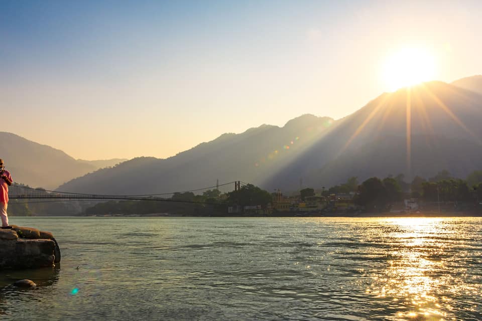 A person standing on a rock by a river with a bridge in the background and mountains in the distance.