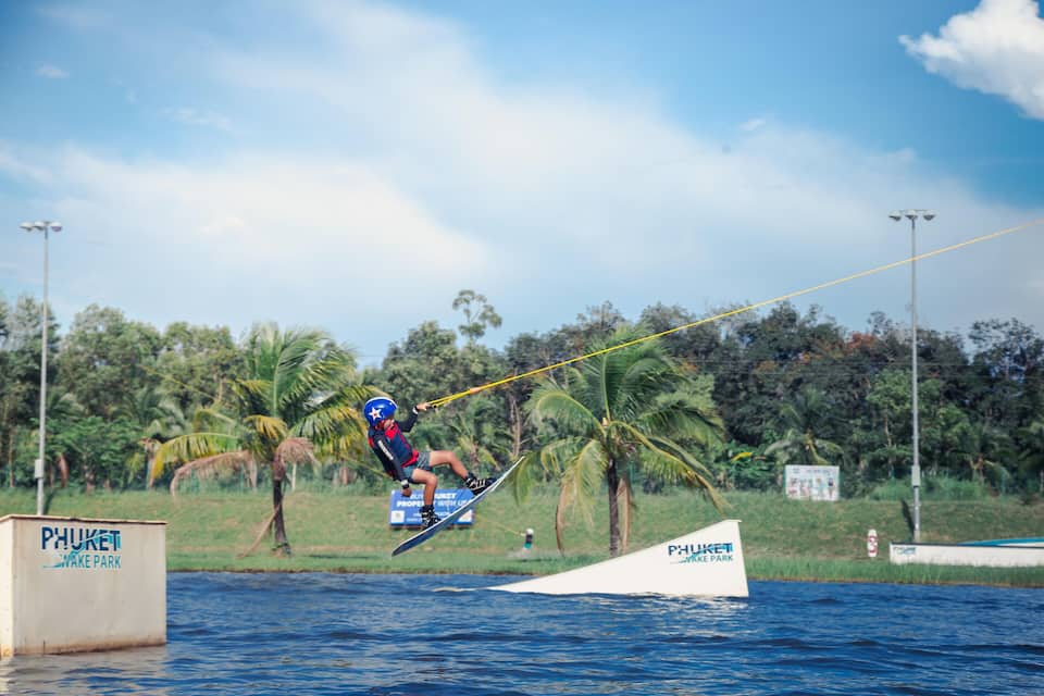Een wakeboarder maakt een sprong over het water, met een wakeboardpark bord op de achtergrond.