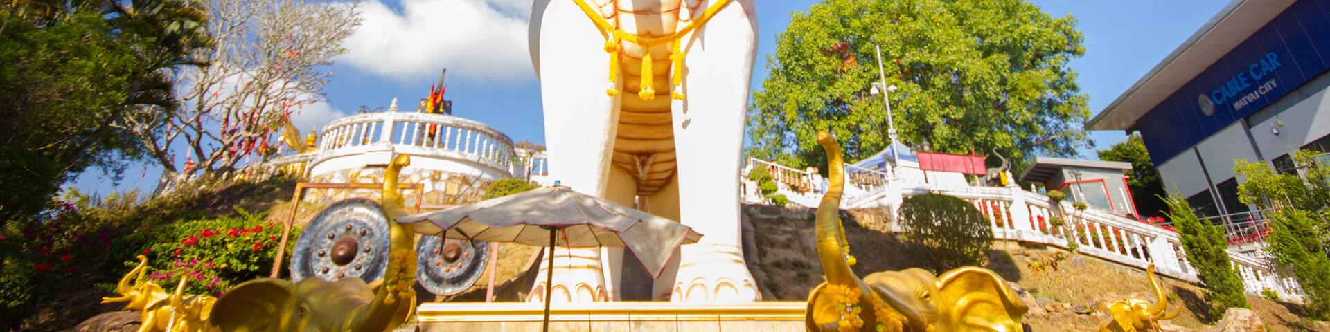 Una gran estatua dorada de elefante sobre un pedestal blanco, rodeada de follaje y un edificio que dice "Cable Car International Center".