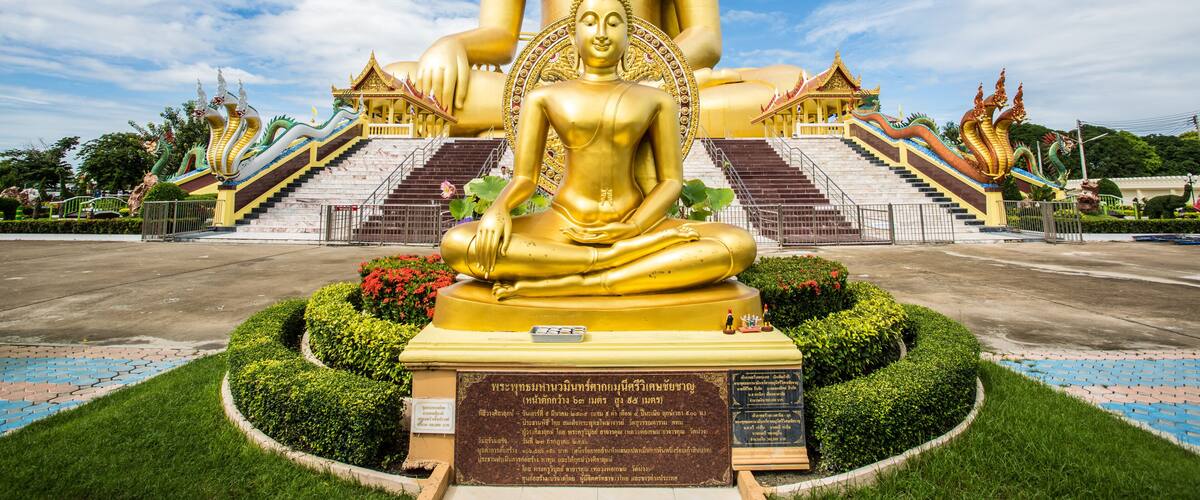 A large golden Buddha statue with a smaller figure in front, surrounded by a temple complex with stairs and traditional architecture.