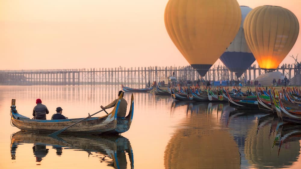 A calm river with traditional boats and hot air balloons in the background.