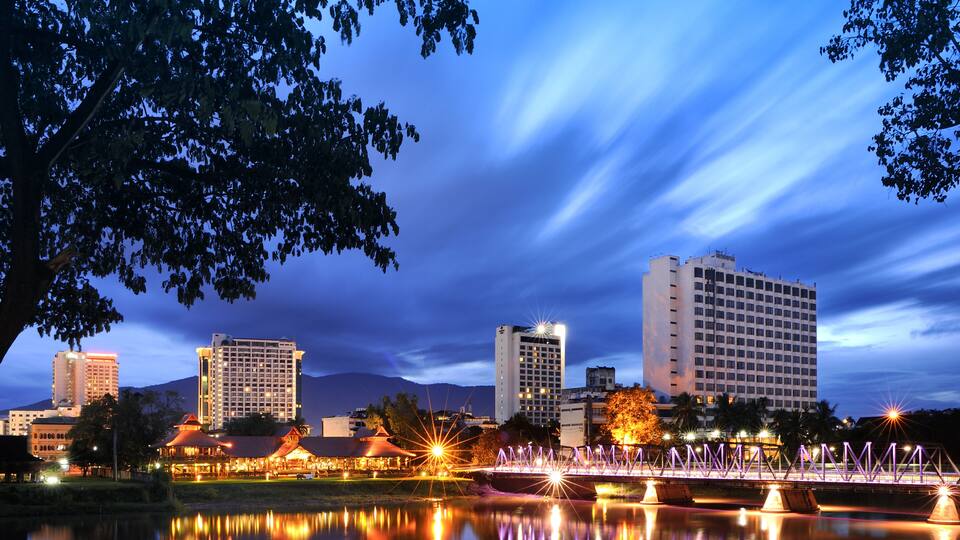 A cityscape at dusk with illuminated buildings, a bridge, and a calm river reflecting the lights.