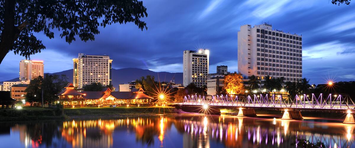 A cityscape at dusk with illuminated buildings, a bridge, and a calm river reflecting the lights.