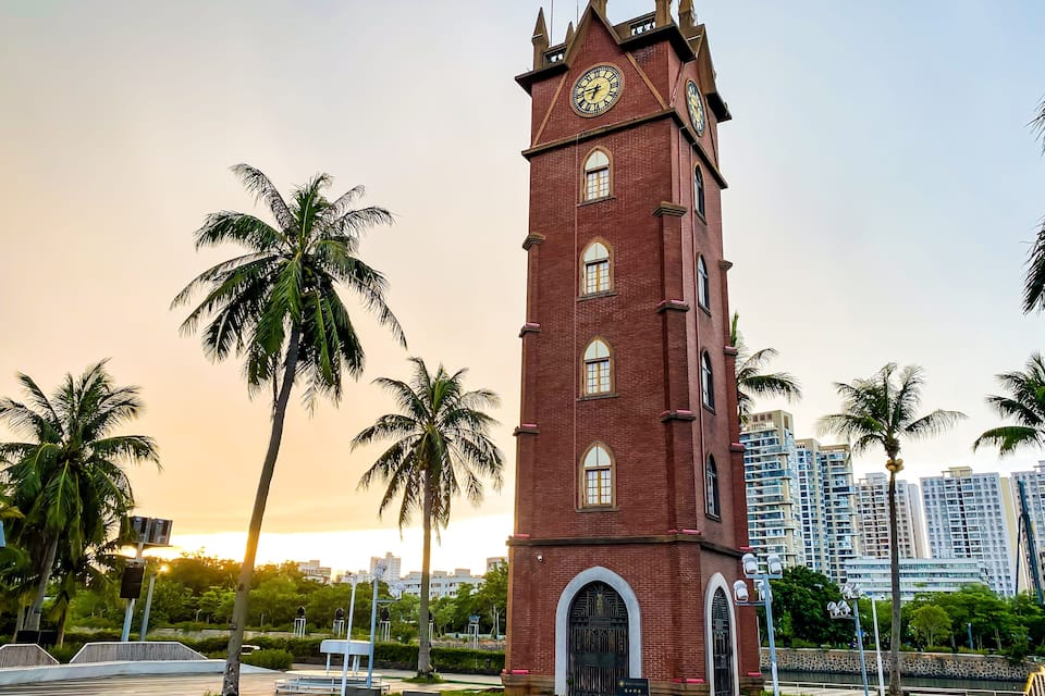 Una torre de reloj de ladrillo rojo con esfera horaria, rodeada de palmeras y edificios modernos.