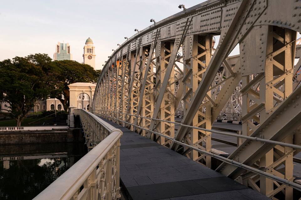 Un puente de acero con un paseo de piedra, una torre del reloj y un edificio con ventanas de arco en el fondo.