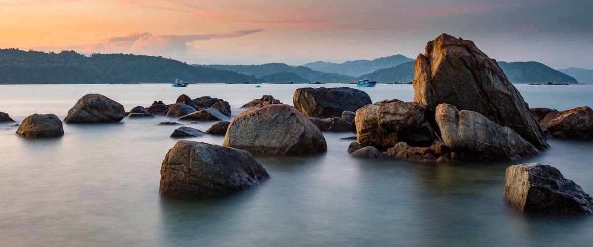 Large boulders in a calm body of water with boats in the distance and mountains in the background.