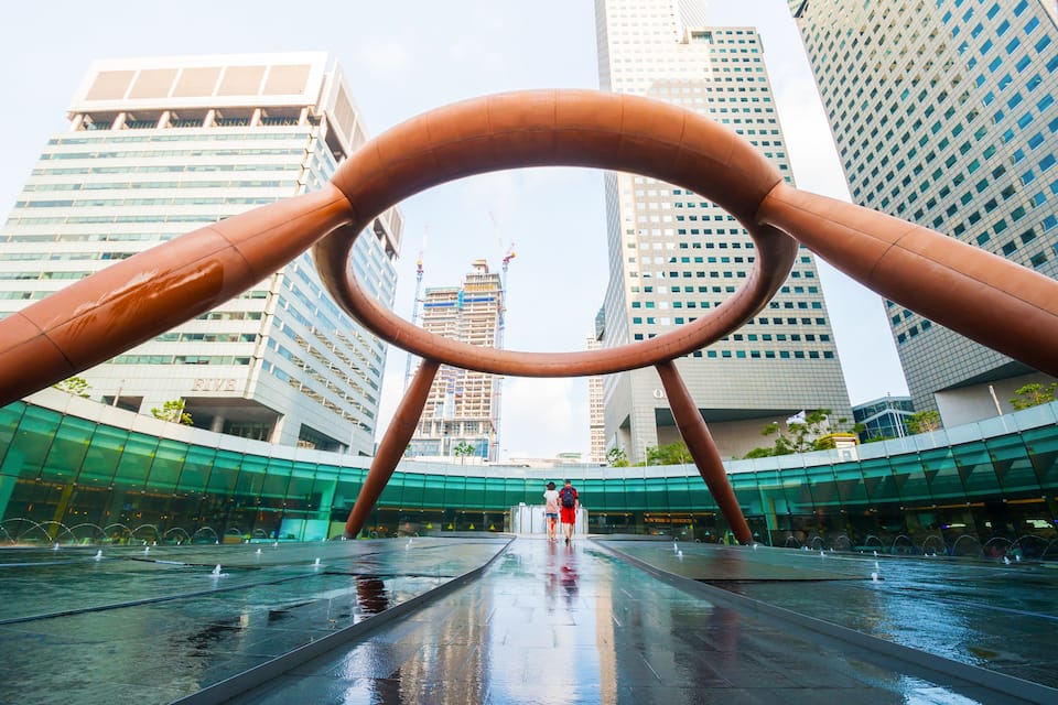 A modern urban plaza with a large circular sculpture, surrounded by high-rise buildings and a wet reflective surface.