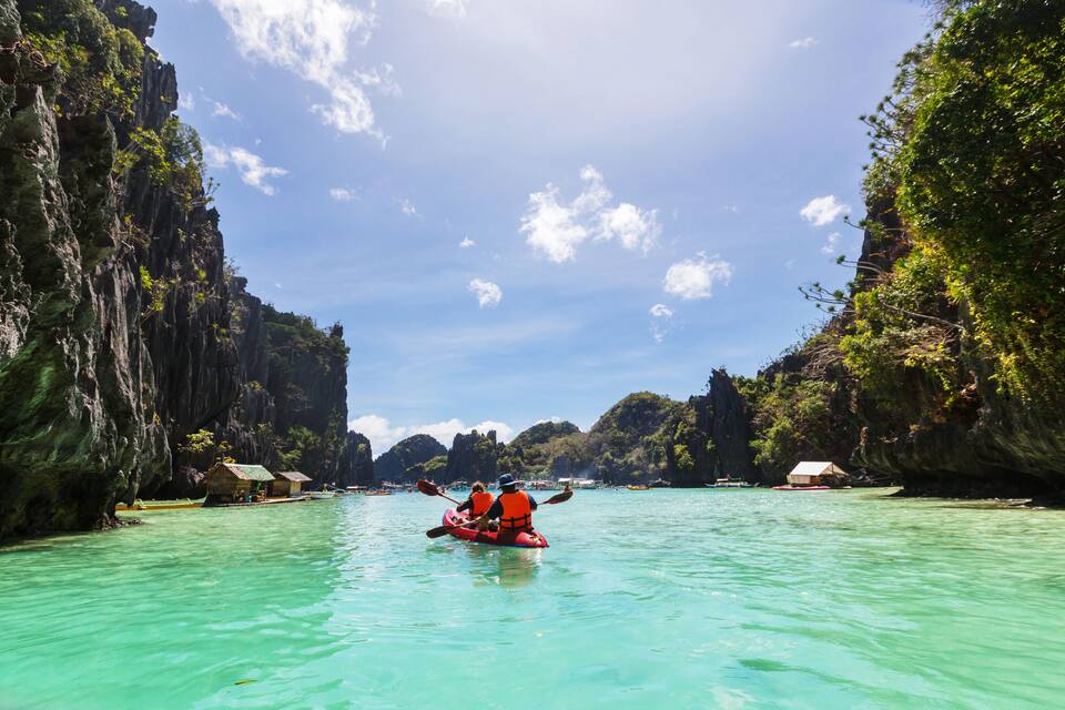 A clear turquoise sea with a kayak in the middle, surrounded by rocky cliffs and small huts.