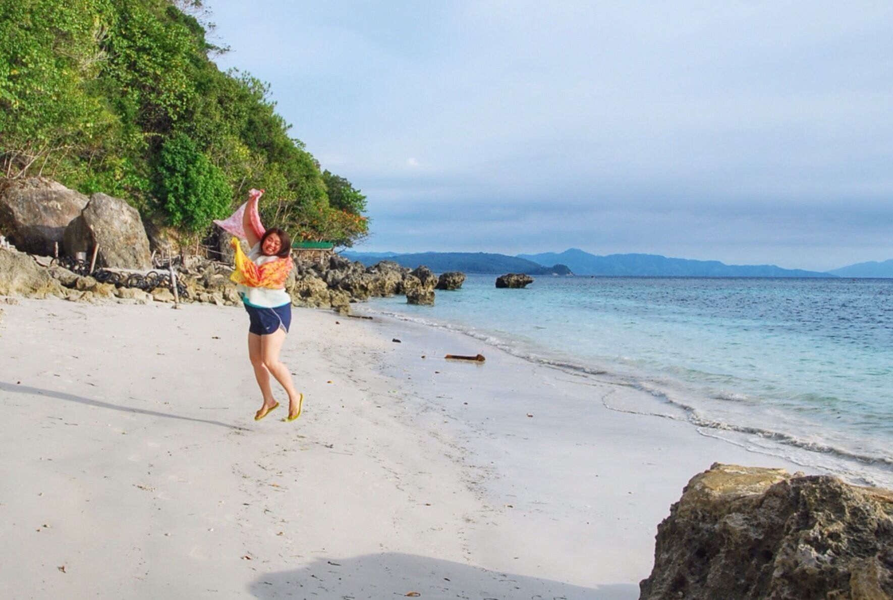 A beach with clear water, rocks, and a hilly landscape in the background.