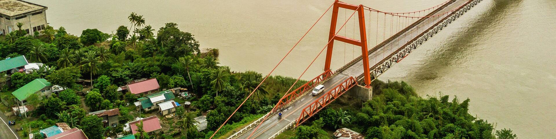 Magapit Suspension Bridge and Cagayan River, largest by volume and longest river in the Philippines. Aerial shot of wide silty river and red bridge.