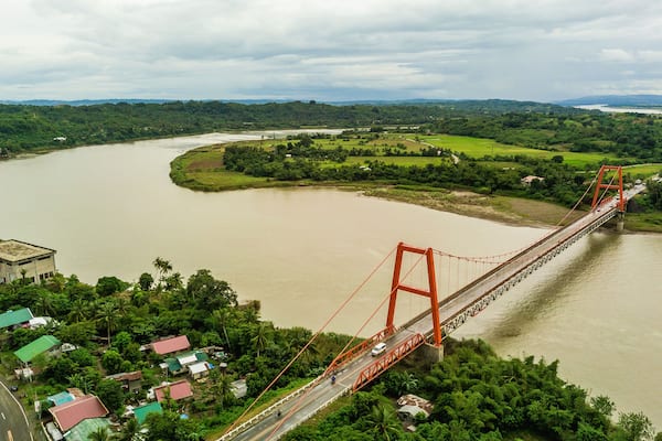 Magapit Suspension Bridge and Cagayan River, largest by volume and longest river in the Philippines. Aerial shot of wide silty river and red bridge.