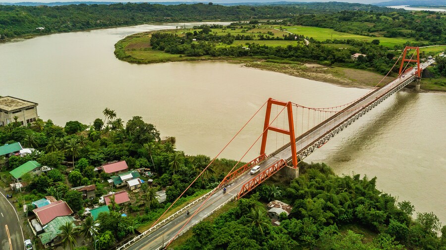 Magapit Suspension Bridge and Cagayan River, largest by volume and longest river in the Philippines. Aerial shot of wide silty river and red bridge.