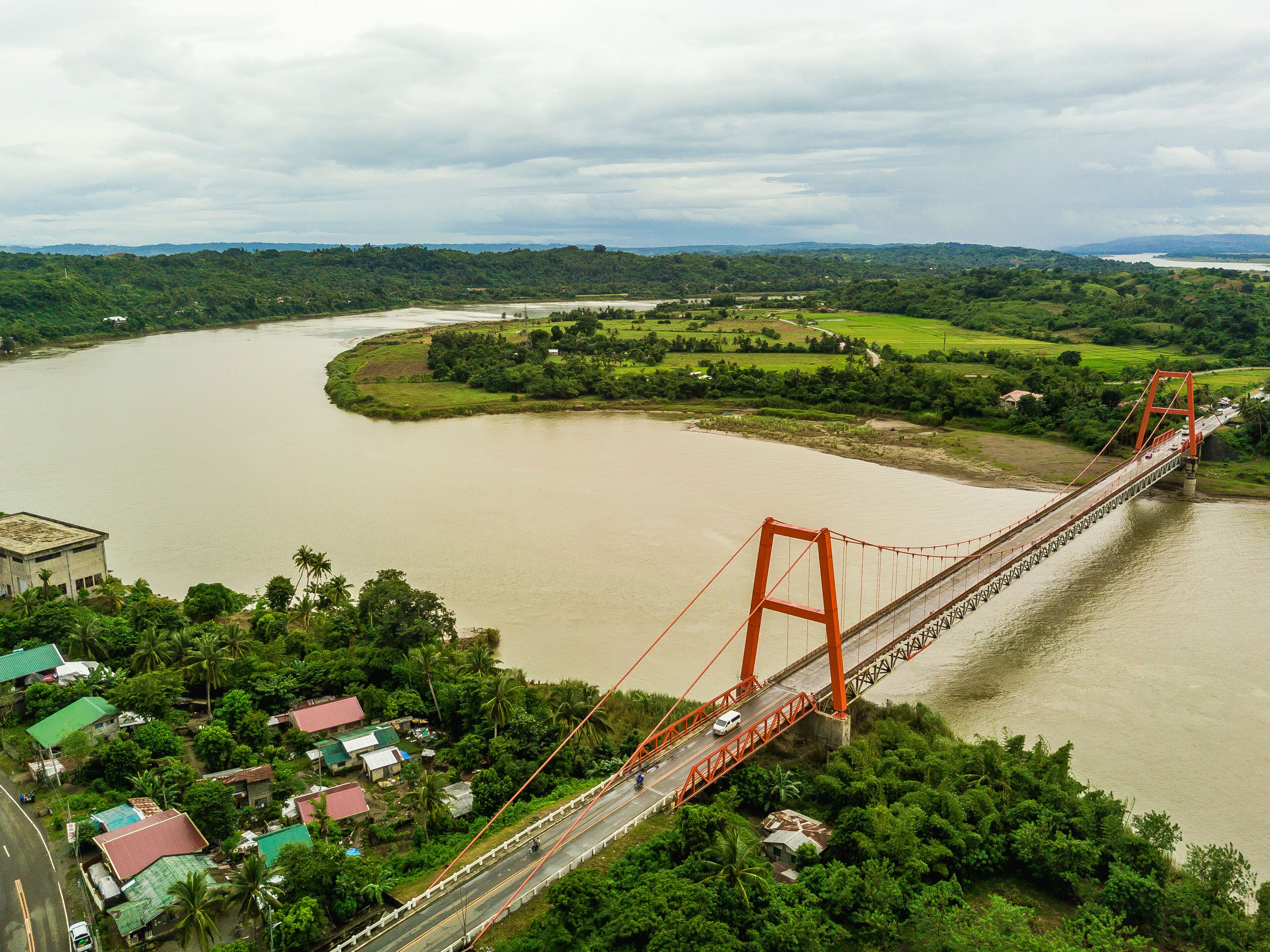 A large orange bridge spanning a wide river, surrounded by lush greenery and distant hills.