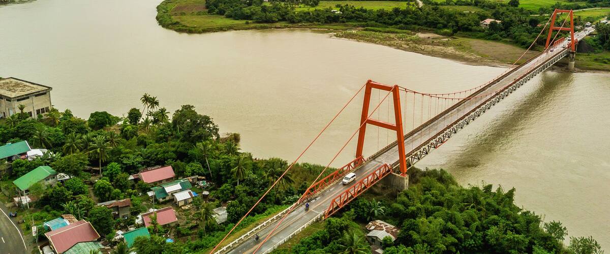 A large orange bridge spanning a wide river, surrounded by lush greenery and distant hills.