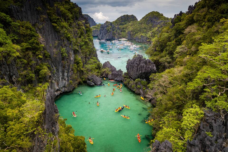 A turquoise bay surrounded by steep cliffs, with several kayaks and boats anchored in the water.