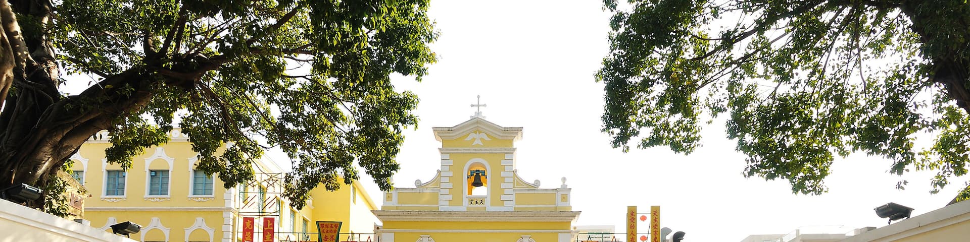 A yellow building with a bell tower, surrounded by a cobblestone courtyard with arches and trees.