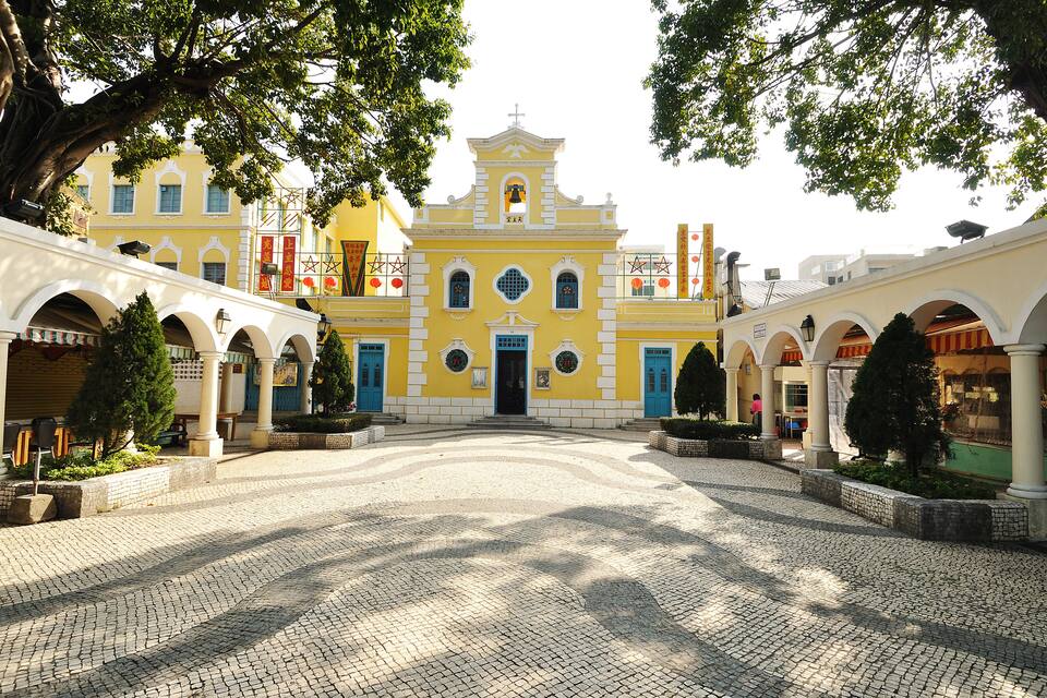 A yellow building with a bell tower, surrounded by a cobblestone courtyard with arches and trees.