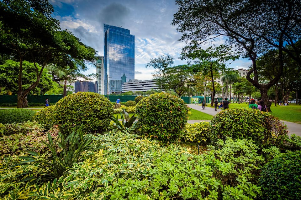 Um parque com arbustos bem cuidados, árvores e um arranha-céu imponente ao fundo.