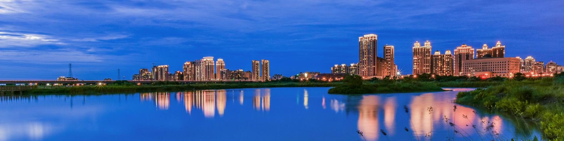 A city skyline at dusk with reflections in a calm body of water, surrounded by greenery.