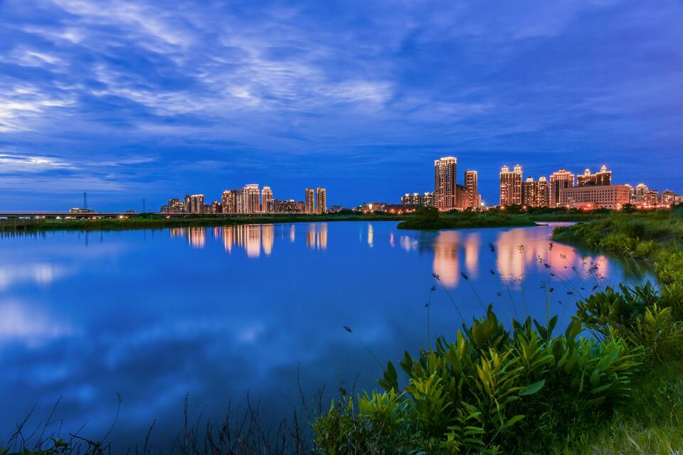 A city skyline at dusk with reflections in a calm body of water, surrounded by greenery.