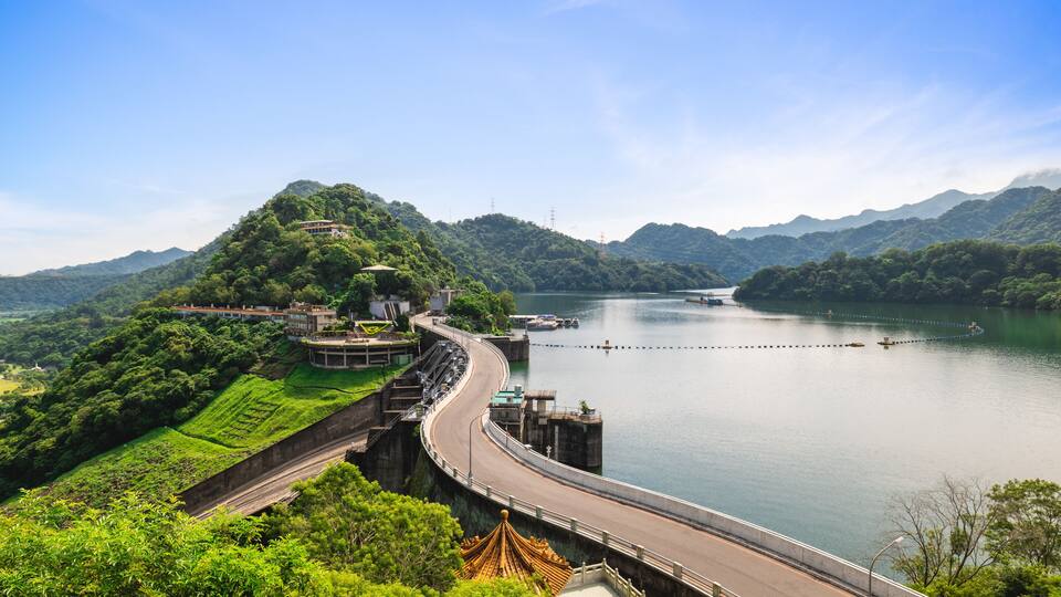 Uma ponte curva sobre um lago tranquilo, rodeada por colinas verdes exuberantes e um céu azul límpido.