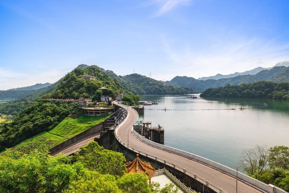 A bridge curving over a calm lake, surrounded by lush green hills and a clear blue sky.