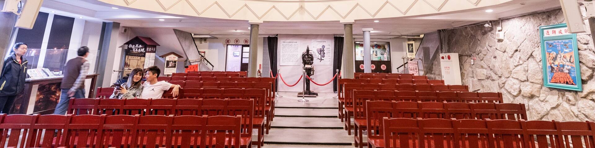 Un auditorio interior con sillas rojas, un escenario y una pared de piedra con carteles.