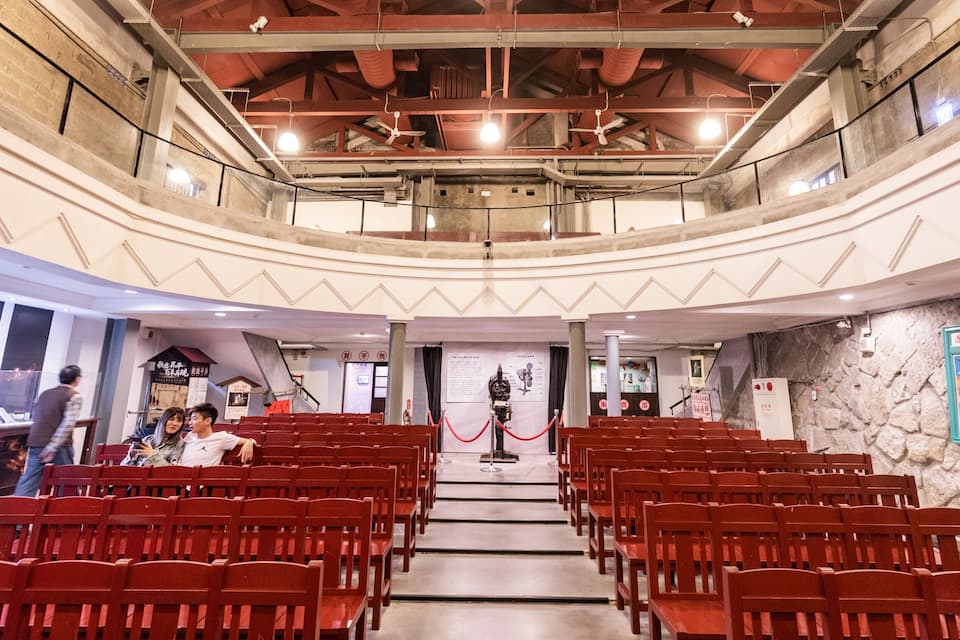 An indoor auditorium with red chairs, a stage, and a stone wall with posters.