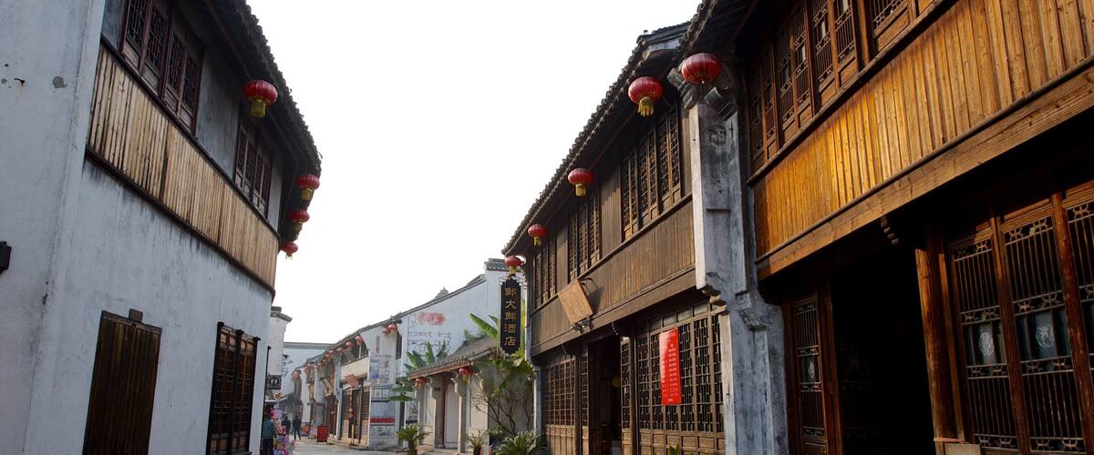 A narrow street lined with traditional wooden buildings, red lanterns, and potted plants.