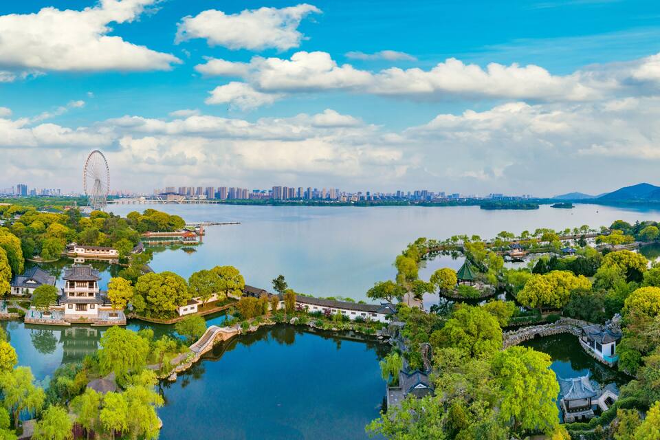 A scenic lakeside park with traditional architecture, a Ferris wheel, and a city skyline in the distance.