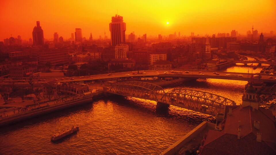 Eine Stadtansicht mit einer markanten Brücke über einem Fluss, umgeben von verschiedenen Gebäuden und einem Sonnenuntergang.