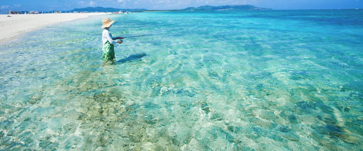 A person wading in clear turquoise waters near a sandy beach with distant mountains.