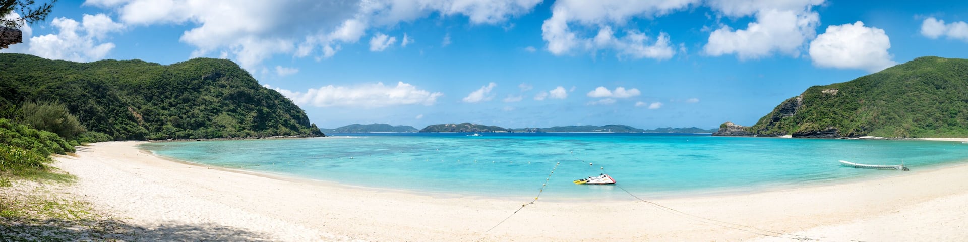 A beach with clear turquoise water, a boat anchored in the middle, and lush green hills in the background.