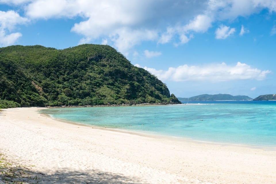 A beach with clear turquoise water, a boat anchored in the middle, and lush green hills in the background.