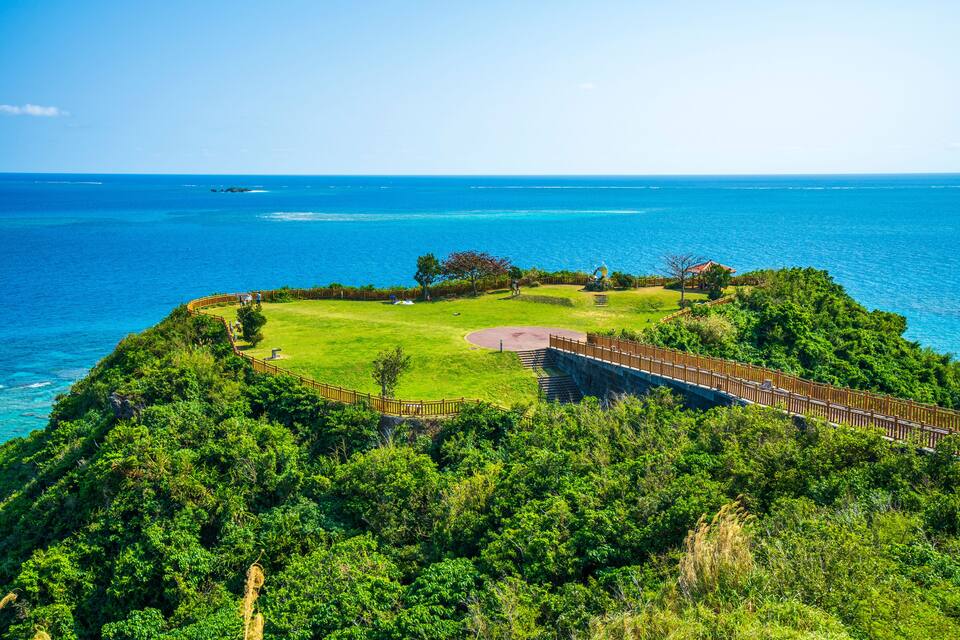 A coastal landscape with a grassy area, a wooden fence, and a clear view of the ocean.